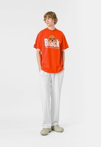 Camiseta gráfica roja con el texto "Black" y un diseño de oso, combinada con pantalones anchos blancos y calzado verde tipo slip-on.