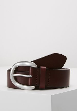 Tummanruskea nahkavyö kiedottuna valkoiselle pinnalle, suuri pyöreä hopeanvärinen metallisolki näkyvissä edessä.