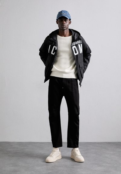 Black windbreaker with large white lettering, cream knit sweater, black pants, and beige sneakers. Denim cap completes the outfit.