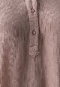 Tessuto ruvido di un leggero rosa pallido con una patta di bottoni che presenta due bottoni coordinati. La texture è morbida e increspata, con un orlo angolato.