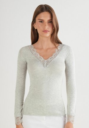 Kobieta ubrana w jasnoszarą bluzkę z długim rękawem, z koronkową lamówką na dekolcie w kształcie litery V oraz na mankietach, stojąca na jednolitym białym tle.