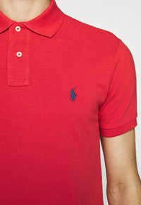 Personne portant un polo rouge avec deux boutons et un petit logo brodé de cheval bleu sur la poitrine gauche.