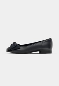 Chaussure plate noire avec un petit talon et un grand nœud en tissu à l’avant, montrée de profil sur un fond uni.