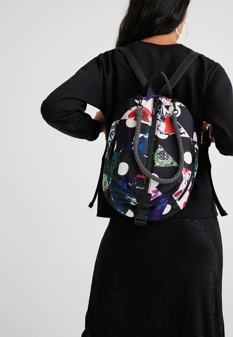 Femme portant un sweat-shirt noir et une jupe avec un sac à dos coloré à motif avec des pois blancs et un détail en corde tressée.