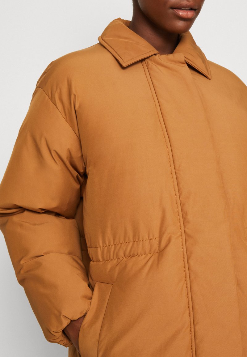 Braune gepolsterte Jacke mit Kragen, ausgestattet mit Seitentaschen und genähten Details. Das Material wirkt glatt und leicht glänzend.