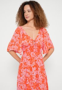 Ziedu raksta kleita ar dziļu V-veida izgriezumu, īsām plandošām piedurknēm un auklu jostu viduklī. Oranža fona krāsa ar rozā un baltu ziedu rakstiem.