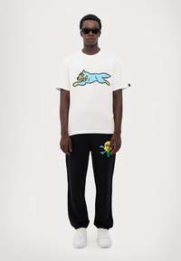 White short-sleeve T-shirt featuring a blue cartoon cat design; paired with black sweatpants displaying a colorful ice cream graphic. White sneakers.