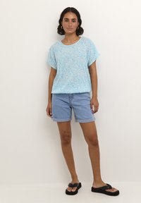 Maglietta azzurro chiaro a maniche corte con un motivo floreale, abbinata a dei pantaloni corti in denim con risvolto. Il modello indossa ciabatte nere.