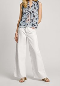 Kvinna iklädd vita vida byxor, ärmlös blå blommig blus och guldiga klacksandaler med remmar, står med en hand i fickan.