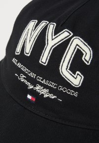 Black baseball cap with white embroidered "NYC" text, "ALL AMERICAN CLASSIC GOODS," and "Tommy Hilfiger" logo. Features small red, white, and blue accent.