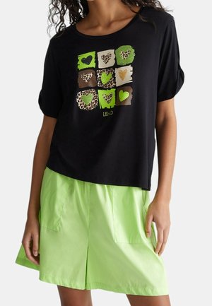 Femme portant un T-shirt noir avec des cœurs verts et imprimé léopard en grille, assorti à un short vert lime vif avec poches avant.