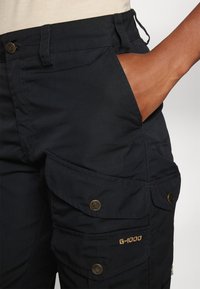 Black cargo pants with a textured fabric, featuring side pockets, brass buttons, and a gold “G-1000” logo detail on the thigh.