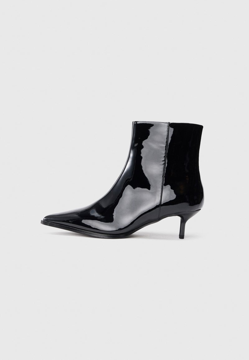 Bottine en cuir verni noir brillant avec un bout pointu et un petit talon aiguille sur un fond uni gris clair.