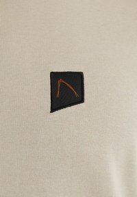 Beige geripptes T-Shirt mit einem schwarzen Patch, der ein orangefarbenes gesticktes geometrisches Design zeigt. Das Detail betont die Textur und den Kontrast.