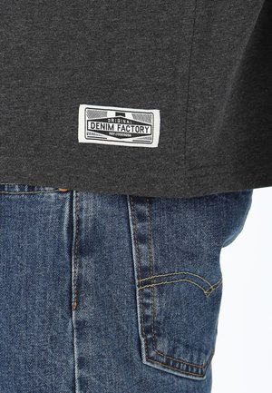 Graues T-Shirt mit einem weißen Etikett mit der Aufschrift "ORIGINAL DENIM FACTORY" am linken Saum, kombiniert mit blauen Jeans im Denim-Look mit gelber Steppnaht.