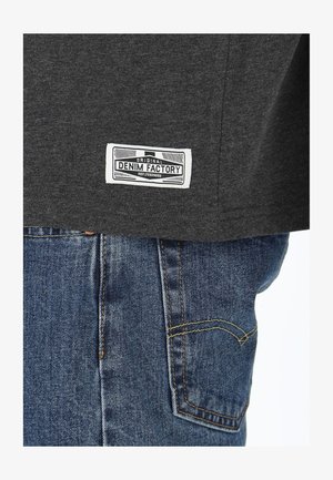 Grå T-shirt med en hvid label der står "ORIGINAL DENIM FACTORY" på venstre søm, parret med blå denim jeans med gule stikninger.