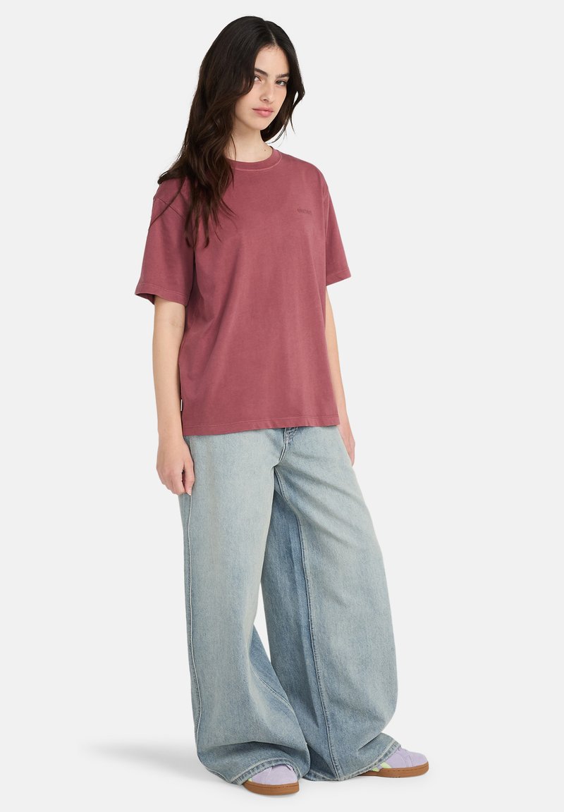 Junge Frau mit langen dunklen Haaren, die ein lockeres mauvefarbenes T-Shirt, weite hellblaue Jeans und lavendelfarbene Turnschuhe vor weißem Hintergrund trägt.