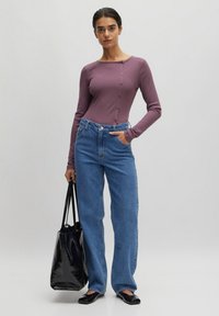 Beerenfarbener Langarmoberteil mit Knopfdetails, kombiniert mit hochgeschnittenen blauen Jeans. Das Model hält eine schwarze Tragetasche und trägt schwarze Ballerinas.