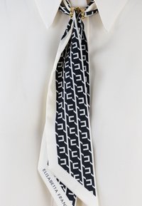 Sciarpa di seta con un motivo geometrico in bianco e nero, caratterizzata da una texture liscia e annodata al collo con accenti in metallo dorato.