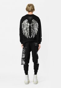 Sudadera negra con gráfico de alas plateadas en la parte posterior, combinada con pantalones jogger negros que presentan detalles en blanco y etiquetas impresas. Botines blancos hasta el tobillo.