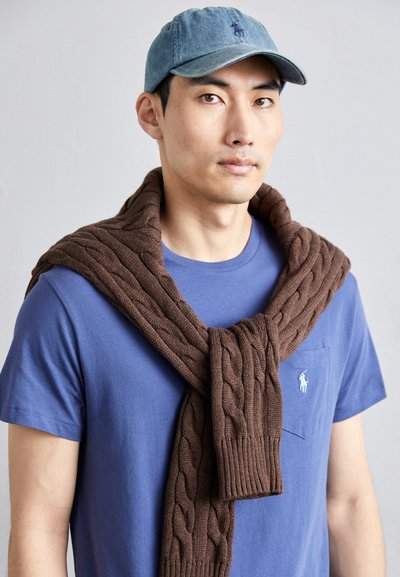 Cardigã de malha castanho atado ao pescoço sobre uma t-shirt azul com um bolso no peito. Boné de ganga azul claro usado na cabeça.