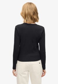 Chemise noire à manches longues avec un col rond, une texture douce et une coupe ajustée, associée à un pantalon de couleur claire et des accents minimalistes.