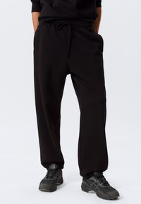 Personne portant un pantalon de survêtement noir à cordon et des chaussures de sport noires, debout les mains dans les poches sur un fond blanc.