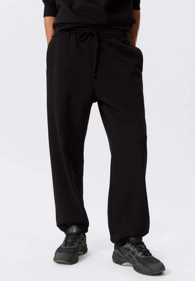 Personne portant un pantalon de survêtement noir à cordon et des chaussures de sport noires, debout les mains dans les poches sur un fond blanc.