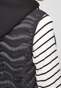 Schwarze Steppweste mit horizontalen genähten Abschnitten, ausgestattet mit einer schwarzen Kapuze und über einem weißen Langarmshirt mit schwarzen horizontalen Streifen getragen.