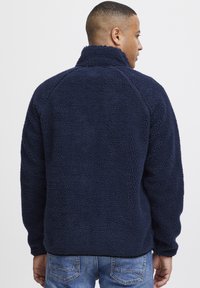 Navy blue fleece pullover with a high collar and textured fabric. Features a relaxed fit and raglan sleeves, paired with blue jeans.
