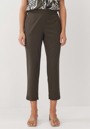 Woman wearing cropped dark olive pants with small front slits paired with strappy beige heeled sandals, standing on white floor.