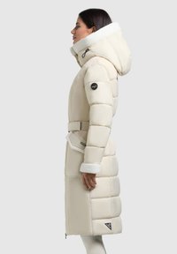 Lange, beige pufferjas met een ceintuur in de taille, een fleece-gevoerde capuchon en contrasterende witte manchetten. Voorzien van een logopatch en ritszakken.