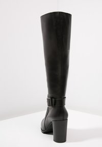 Pitkät mustat nahkasaappaat, joissa on paksu korkea korko ja koristeellinen solkivö kriisitetyssä nilkassa, esitettynä takaa ja sivulta.