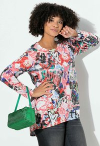 Buntes Langarmhemd mit abstrakten Mustern in Rot, Pink, Blau und Schwarz. Kombiniert mit einer kleinen grünen genarbten Handtasche und dunklen Jeans.