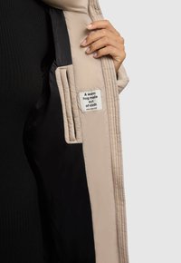 Bež prešit plašč s črnim podlogom, na katerem je etiketa z napisom "Topel objem iz tkanine." Vidna roka, ki drži plašč.