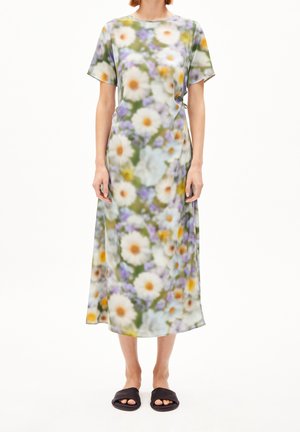 Femme portant une robe à manches courtes, longue jusqu'aux chevilles, avec un motif floral coloré et des sandales noires ouvertes aux orteils, debout devant un fond blanc.