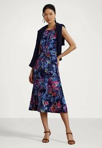 Mulher vestindo um vestido midi sem mangas, floral em tons de azul, com um suéter escuro caído sobre os ombros e sandálias de tiras no tornozelo marrons.