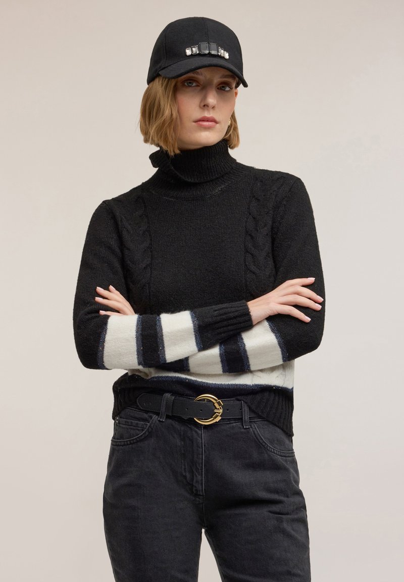 Czarny sweter z wysokim kołnierzem, z detalami w warkoczowy splot oraz kremowo-granatowym paskowanym dołem. W zestawie z czarnymi spodniami i czapką.