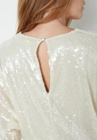 Kremasti top sa sjajnim detaljima i teksturiranim završetkom. Zatvaranje na stražnjoj strani s prorezom i dugmetom. Ima opušteni kroj i sjajnu površinu.