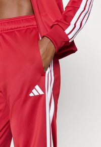 Crvena trenirka s bijelim dizajnom tri pruge na rukavima i nogavici. Ima teksturirani materijal i bijeli Adidas logo.