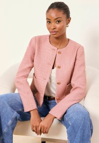 Veste rose texturée à encolure ronde, ornée de quatre boutons dorés, associée à un haut blanc et un jean large bleu clair.