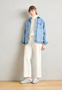 Giacca in denim, blu chiaro, con due tasche sul petto, sopra un maglione spesso color crema. Pantaloni larghi color crema e sneakers blu chiaro con dettagli bianchi.