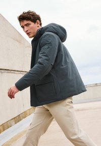 Jachetă impermeabilă de culoare gri închis, cu glugă, având o pungă frontală, textură netedă și o croială lejeră, asortată cu pantaloni bej deschis.