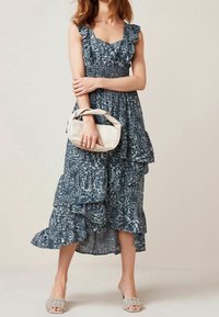 Robe maxi à motifs floraux bleus avec des manches volantées et une jupe superposée. Le mannequin tient un sac à main crème et porte des sandales tressées.