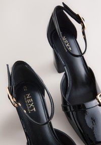 Black patent leather high-heeled shoes with a curved design, adjustable ankle straps, and gold hardware accents. Embossed logo inside.