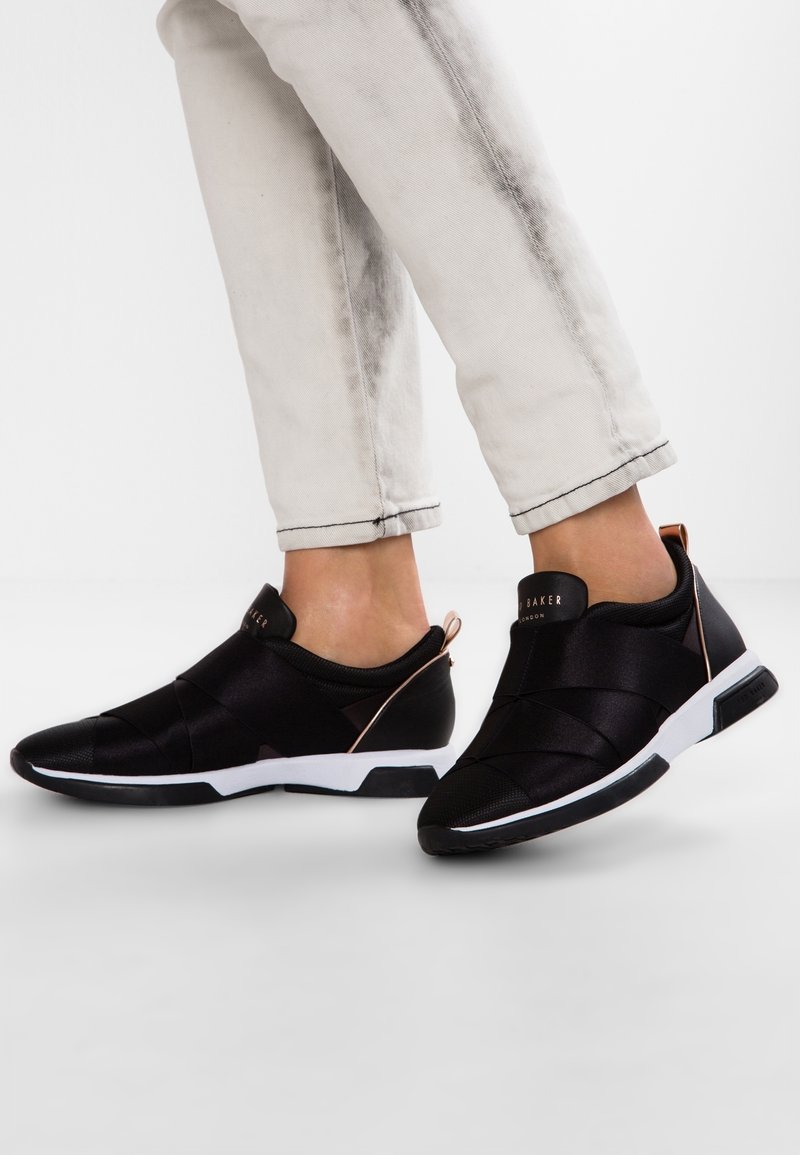 Baskets noires sans lacets avec une tige en tissu texturé, une semelle blanche contrastante et des accents en or rose sur le talon et le logo.