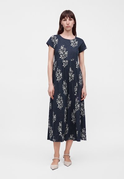 Femme debout portant une robe midi bleu marine avec des imprimés floraux blancs et des manches courtes, associée à des chaussures pointues beige.