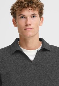 Suéter gris de cuello en v hecho de tejido de punto, con un cuello y una textura sutil. Se lleva sobre una camisa blanca de cuello redondo.