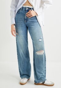 Jeans de mezclilla de pernera ancha en azul claro con detalles desgastados en las rodillas y un cierre de botón negro. Combinados con zapatillas blancas y una camisa desabotonada.