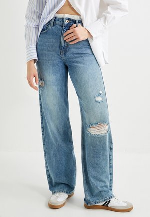 Jeans a gamba larga in denim blu chiaro con dettagli strappati sulle ginocchia e chiusura con bottone nero. Abbinati a sneakers bianche e una camicia sbottonata.